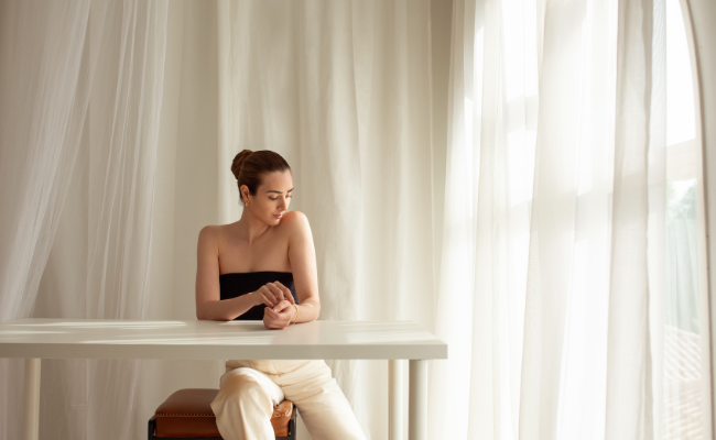 Woman wearing Saint Stones jewellery sitting at a table in a softly lit room with sheer curtains.