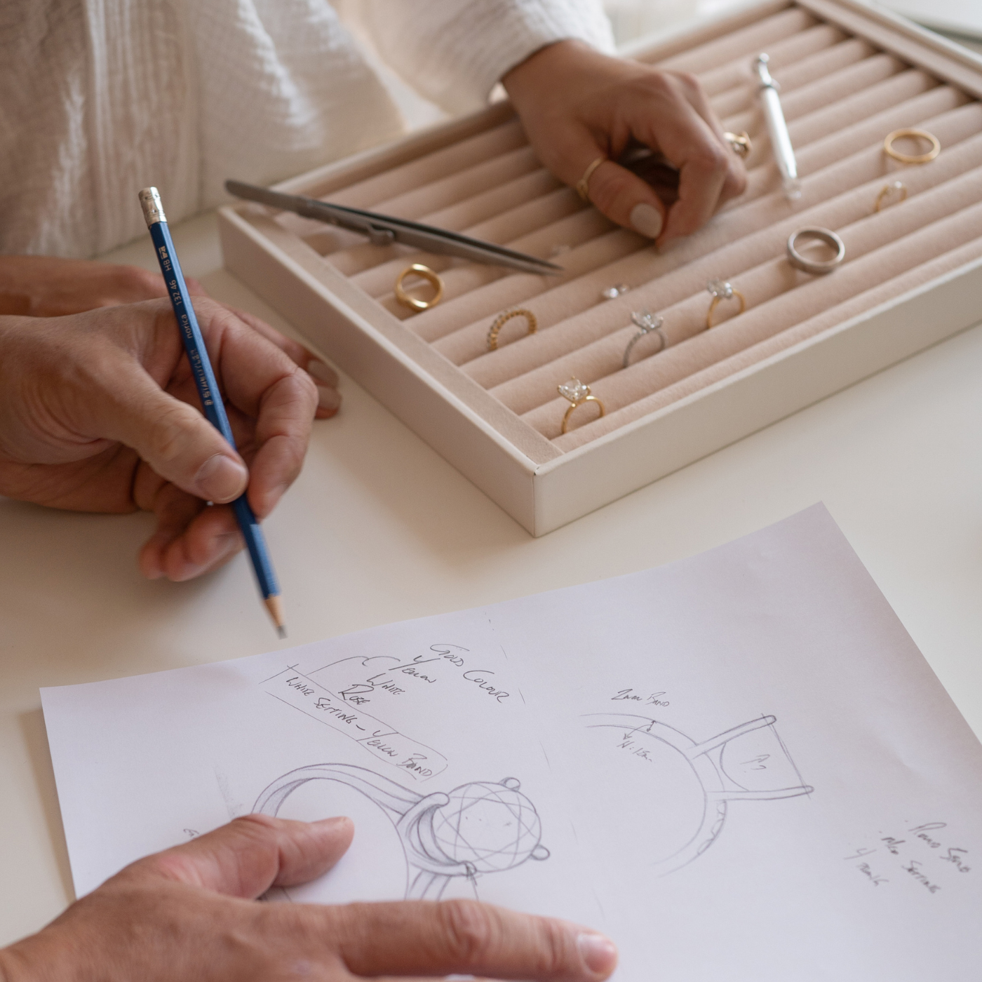 Person drawing jewellery designs on paper with a ring holder displaying wedding and engagement rings by Saint Stones in the background.